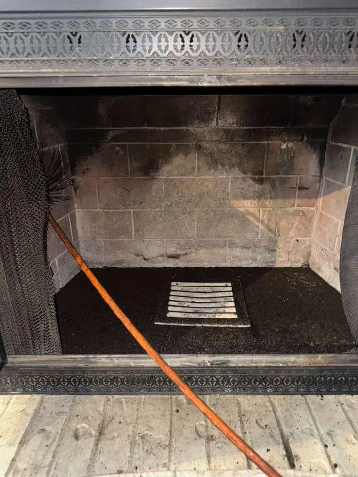 The inside of a firebox under a decorative metal frame, with sooty brick above, a cleared hearth floor with an ash grate and an orange cable lying across it