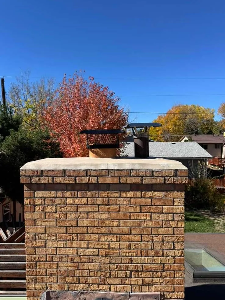 A tan brick chimney with a pale concrete slab on top and two small caps, one a black mesh box, in front of red and gold autumn trees