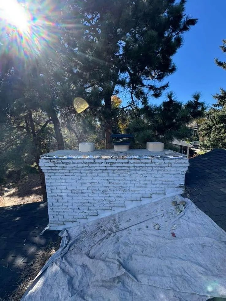 A chimney of white-painted brick with a shiny coated top and a gray tarp spread over the roof below it, with pine trees and sunlit shingles around