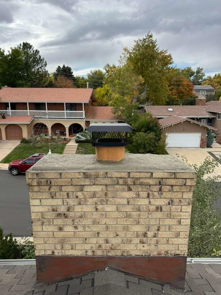 A pale yellow brick chimney with a flat concrete top and a black cap, a dark brown metal band at its base on the roof, above a street of houses and autumn trees
