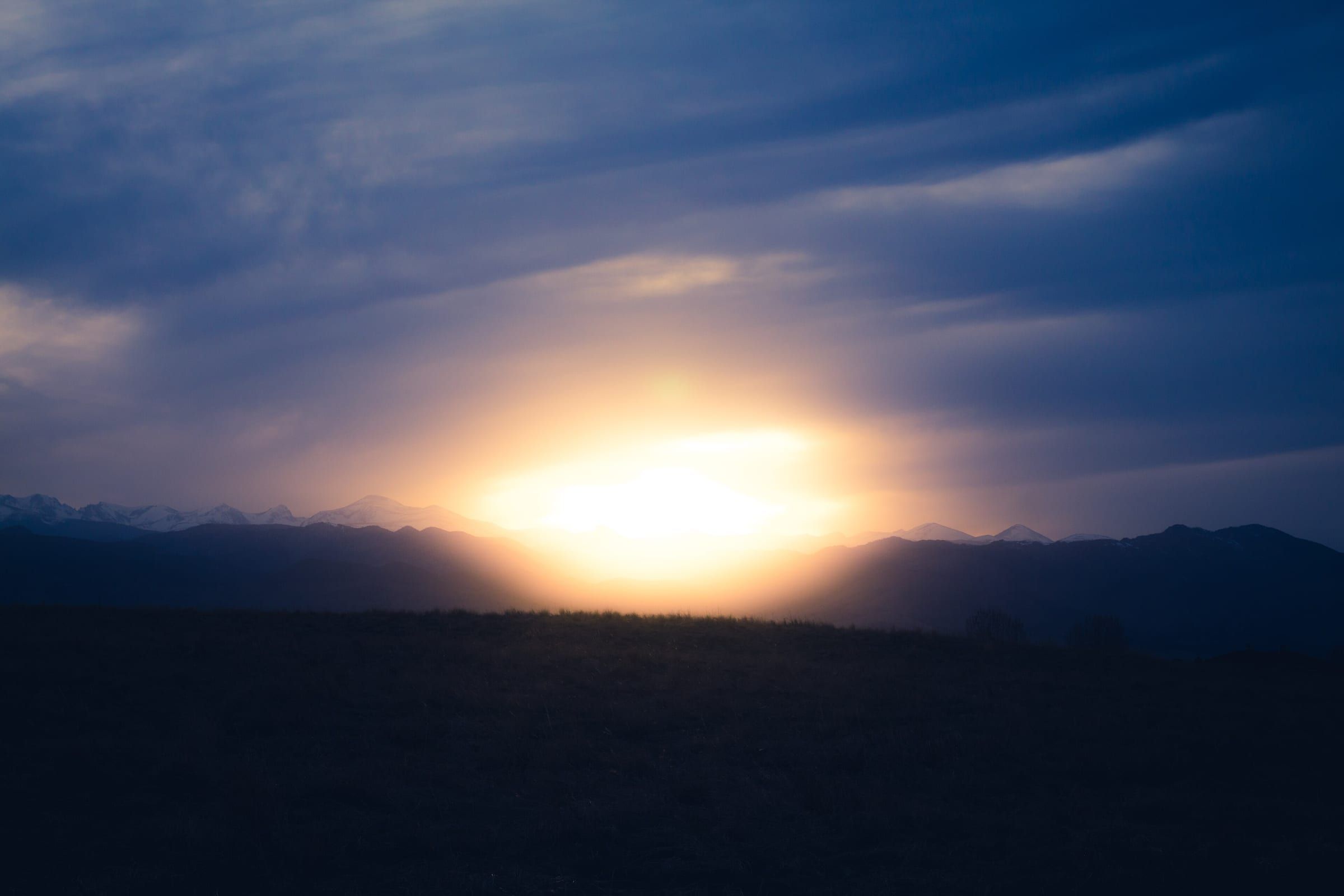 A low sun glowing through layered blue clouds over a dark plain, with a line of mountains, some of them snow-topped, along the horizon